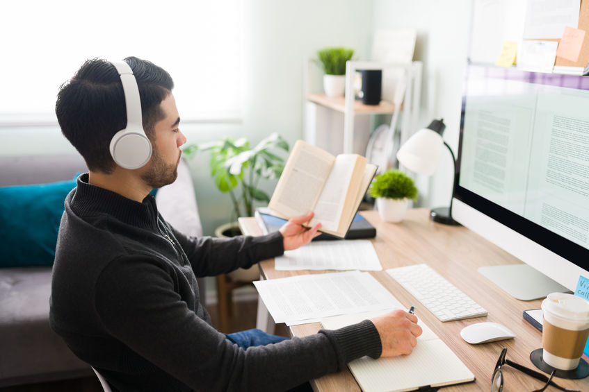 Man in his 20s sitting at his desk at the office and listening to music with headphones.